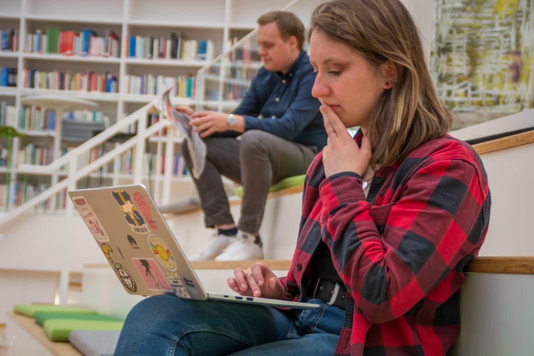 Två studenter som sitter i ett biliotek. Ena studenten surfar på sin laptop medan sen andra läser en tidning.