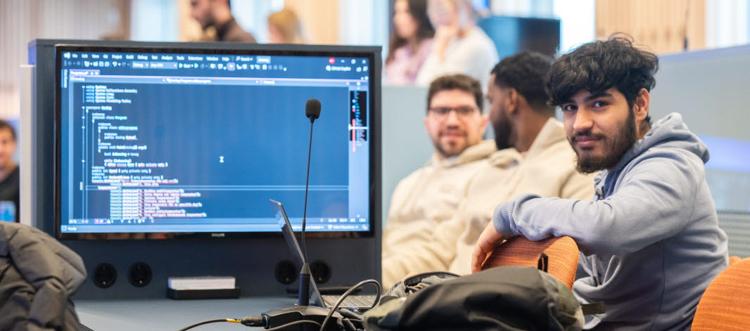 Studenter som sitter i ett klassrum. Fokus ligger på en student som sitter med sin laptop och kollar rakt in i kameran