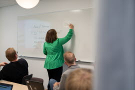 Kvinna står och skriver på whiteboard med ryggen vänd mot åhörarna. 