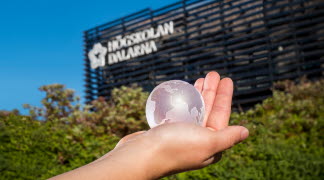 En hand som håller i en glaskula som ser ut som jorden. I bakgrunden gröna buskar och Högskolan Dalarnas fasad.