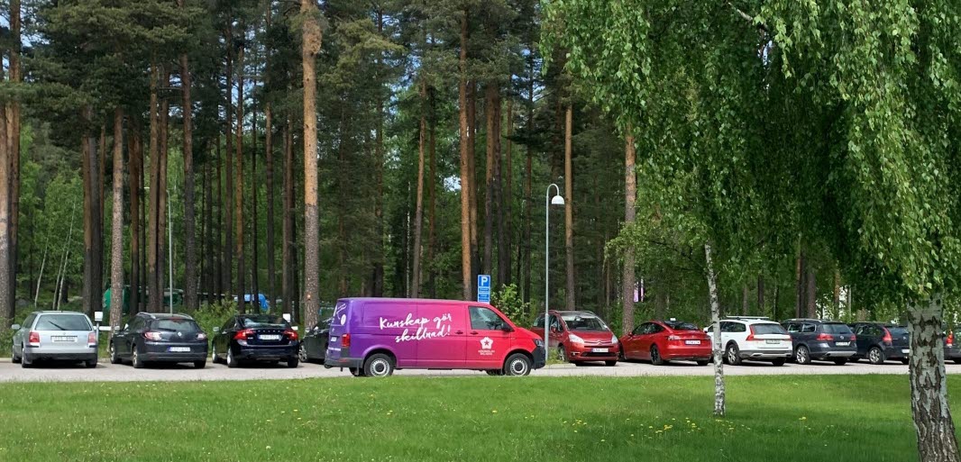 Flera bilar står parkerades, en lila bil med texten Kunskap för skillnad kör på vägen. Det är en grön grasmatta i förgrunden och tallar i bakgrunden.