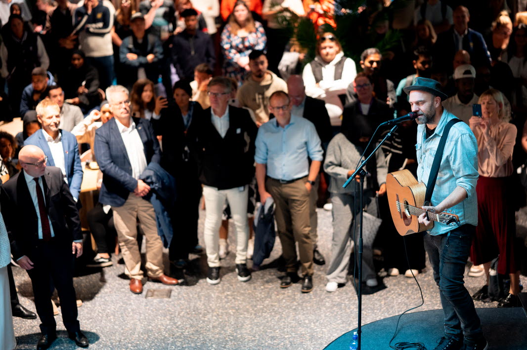 En peson står framför en grupp och spelar gitarr. Personen har hatt. 