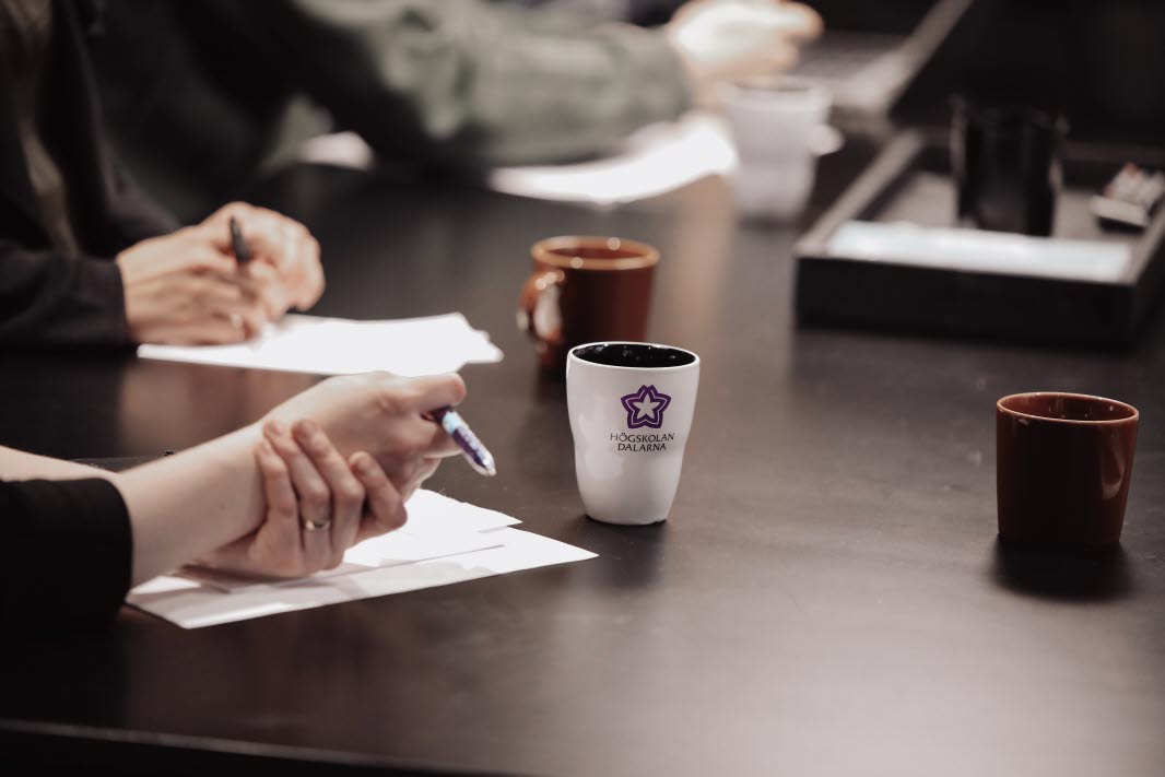 Mug with Högskolan Dalarna logo, table indoor, hands holding pen over paper.