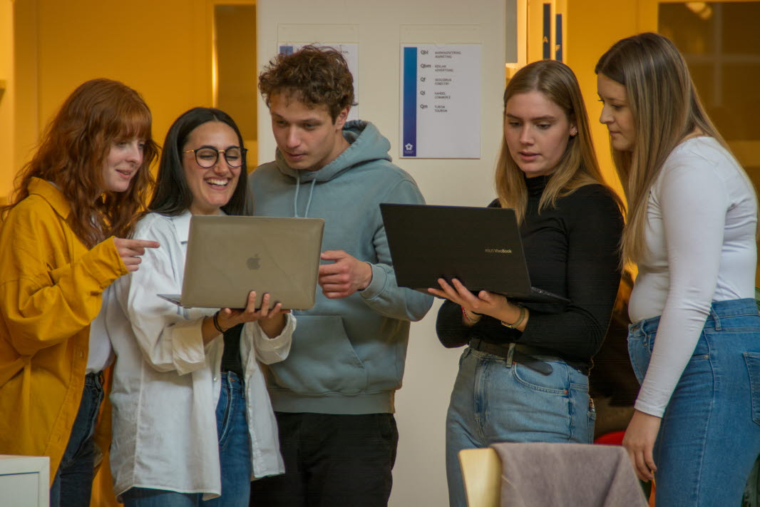 Fem studenter som står upp och tittar på två datorskärmar 