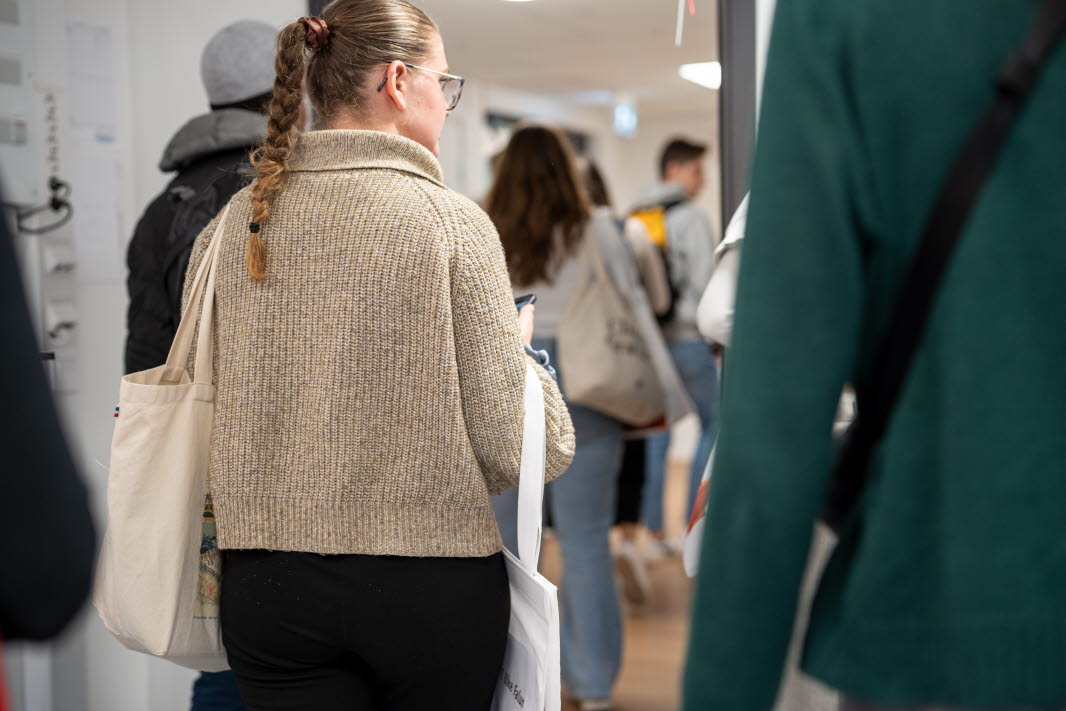 Personer går in i lokal, person med beige tröja och vit tygkasse på axeln.