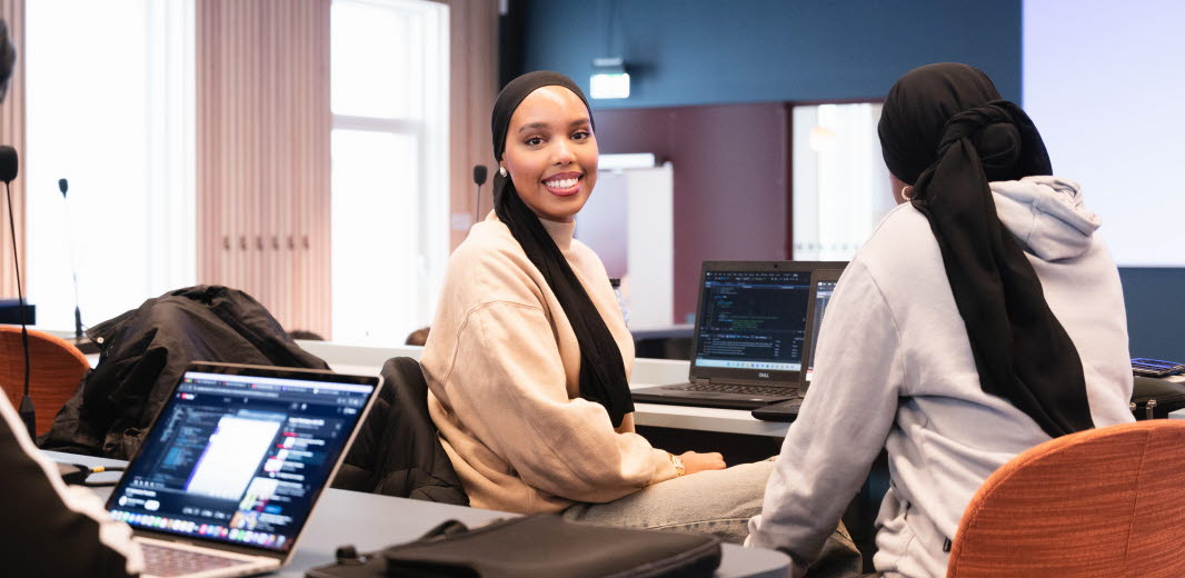 Studenter i ett klassrum som studerar vid sina datorer. En av studenterna kollar rakt in i kameran. 