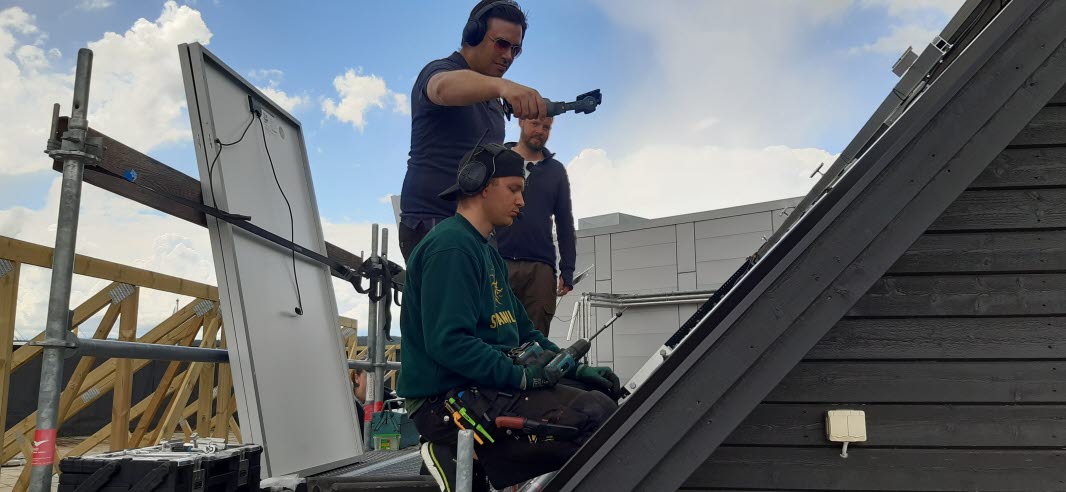 Tre män, en sitter ner på huk och utför ett arbete på ett tak. En av de andra håller i en mickrofon. 