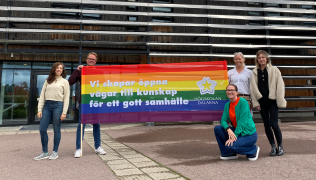 Personer står utomhus håller en banderoll med Pridefärger och texten: Vi skapar öppna vägar till kunskap för ett gott samhälle.