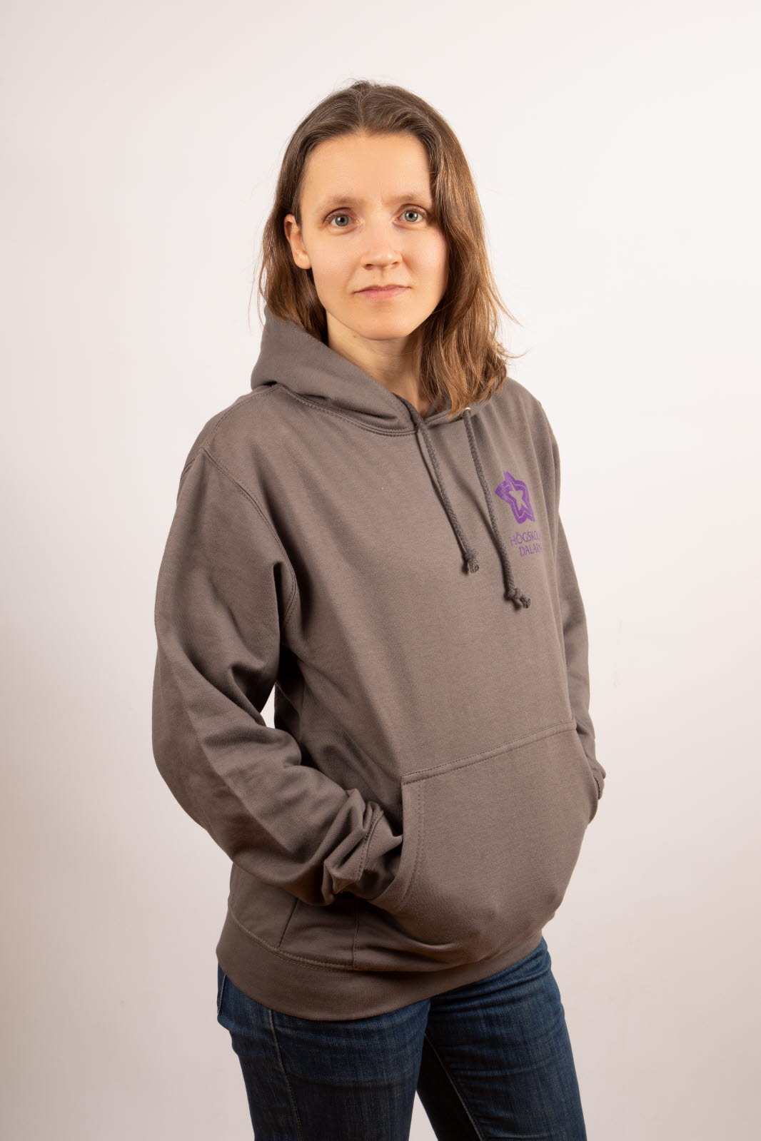 Person i grå huvtröja med Högskolan Dalarnas logga på i lila. Personen har händerna i fickorna och tittar in i kameran.