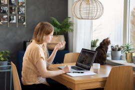 En kvinna sittandes vid ett skrivbord i hemmamiljö med sin laptop. En katt sitter på skrivbordet och tittar ut genom ett fönster.