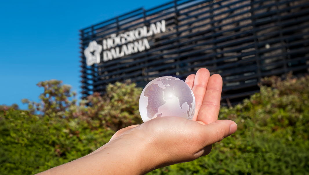 En hand som håller i en glaskula som ser ut som jorden. I bakgrunden gröna buskar och Högskolan Dalarnas fasad.