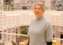Kvinna står inomhus bibliotek grå tröja ljust hår