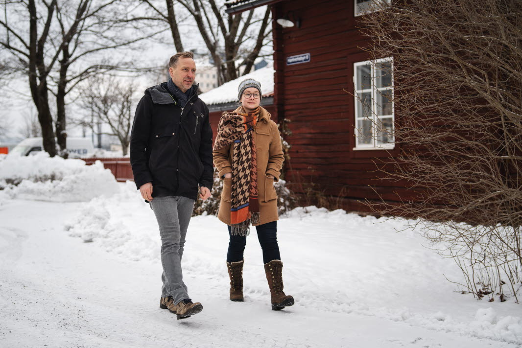 Man i svart jacka och kvinna i brun rock promenerar tillsammans på en gata med snö på backen.